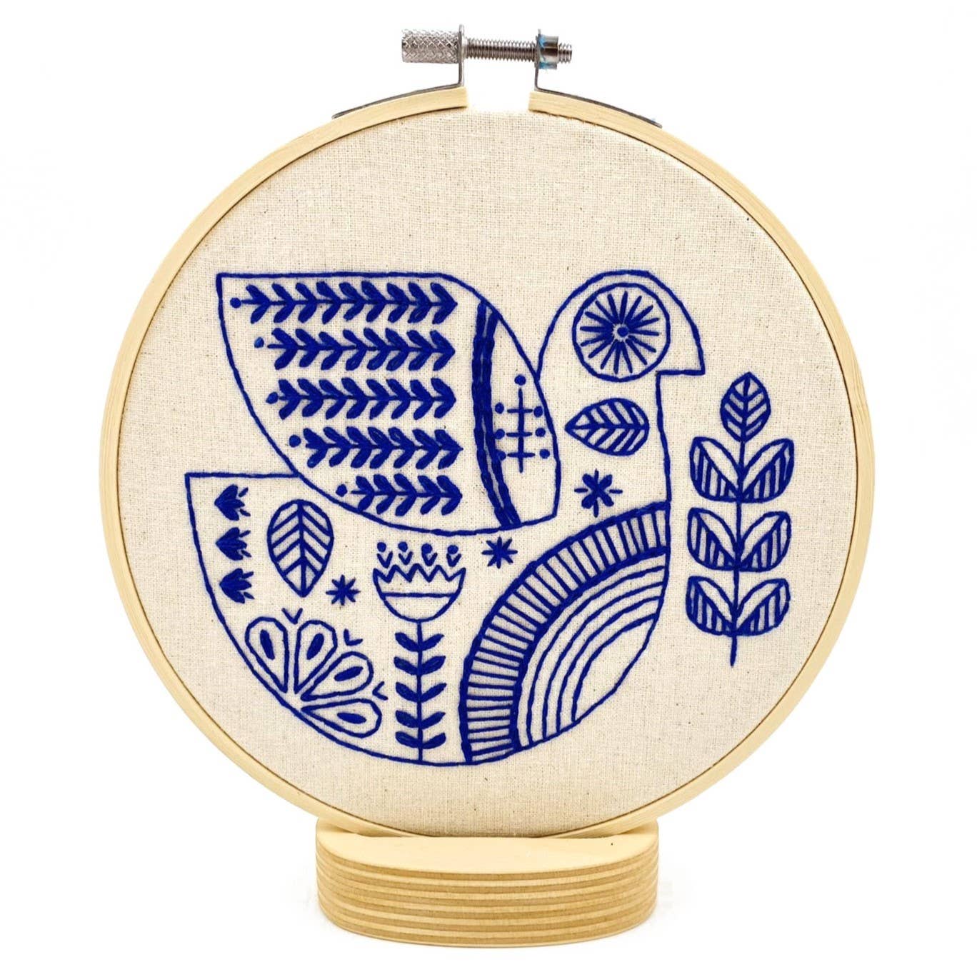The Hygge Dove Complete Embroidery Kit by Hook, Line & Tinker features a blue-stitched stylized bird with geometric patterns and leaves on beige fabric. Includes an upright embroidery hoop on a wooden base, ideal for display.