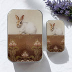 Two small rectangular Firefly Notes "Sweet White Bunny Notions Tin" boxes with vintage-style white rabbit and floral designs on the lids are displayed on a white surface beside a sprig of purple lavender, highlighting their unique handcrafted charm.