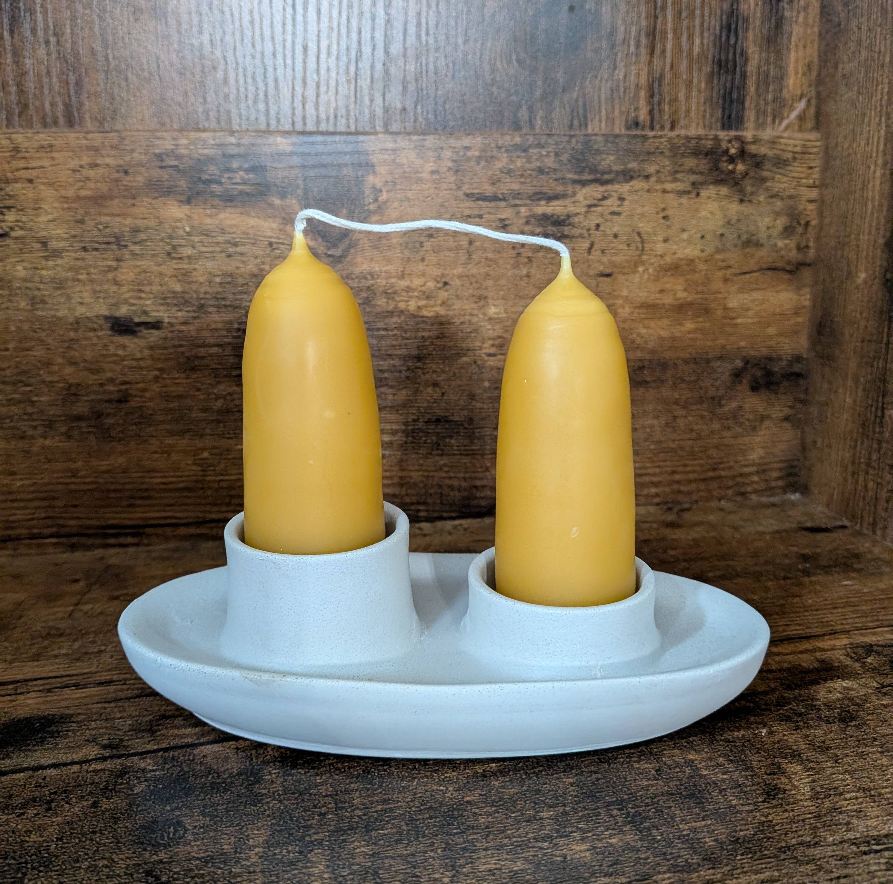 A pair of British-style, hand-dipped Beeswax Stubby Candles by Yarrow Bone Farmstead stand upright in a white ceramic holder with two circular slots, set against a wooden background.