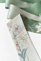 A close-up of Lost Pattern NYC's Lily Silk Twilly Scarf: Green, showcasing elegant white lilies, green leaves, and red accents—perfect for gift wrapping or adding flair as a chic accessory.