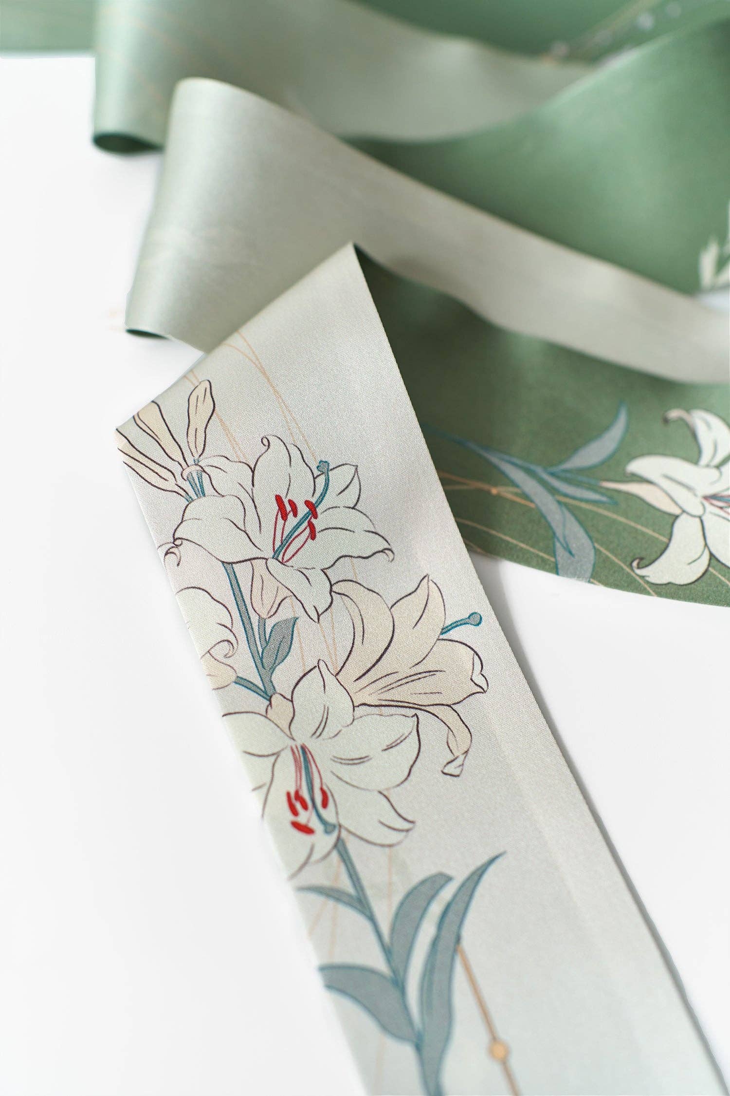 A close-up of Lost Pattern NYC's Lily Silk Twilly Scarf: Green, showcasing elegant white lilies, green leaves, and red accents—perfect for gift wrapping or adding flair as a chic accessory.