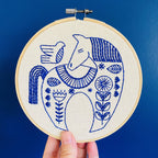 A hand holds up the Hygge Horse Complete Embroidery Kit by Hook, Line & Tinker, featuring a blue and white stylized horse, bird, and floral design stitched on cream fabric, set against a solid blue background.