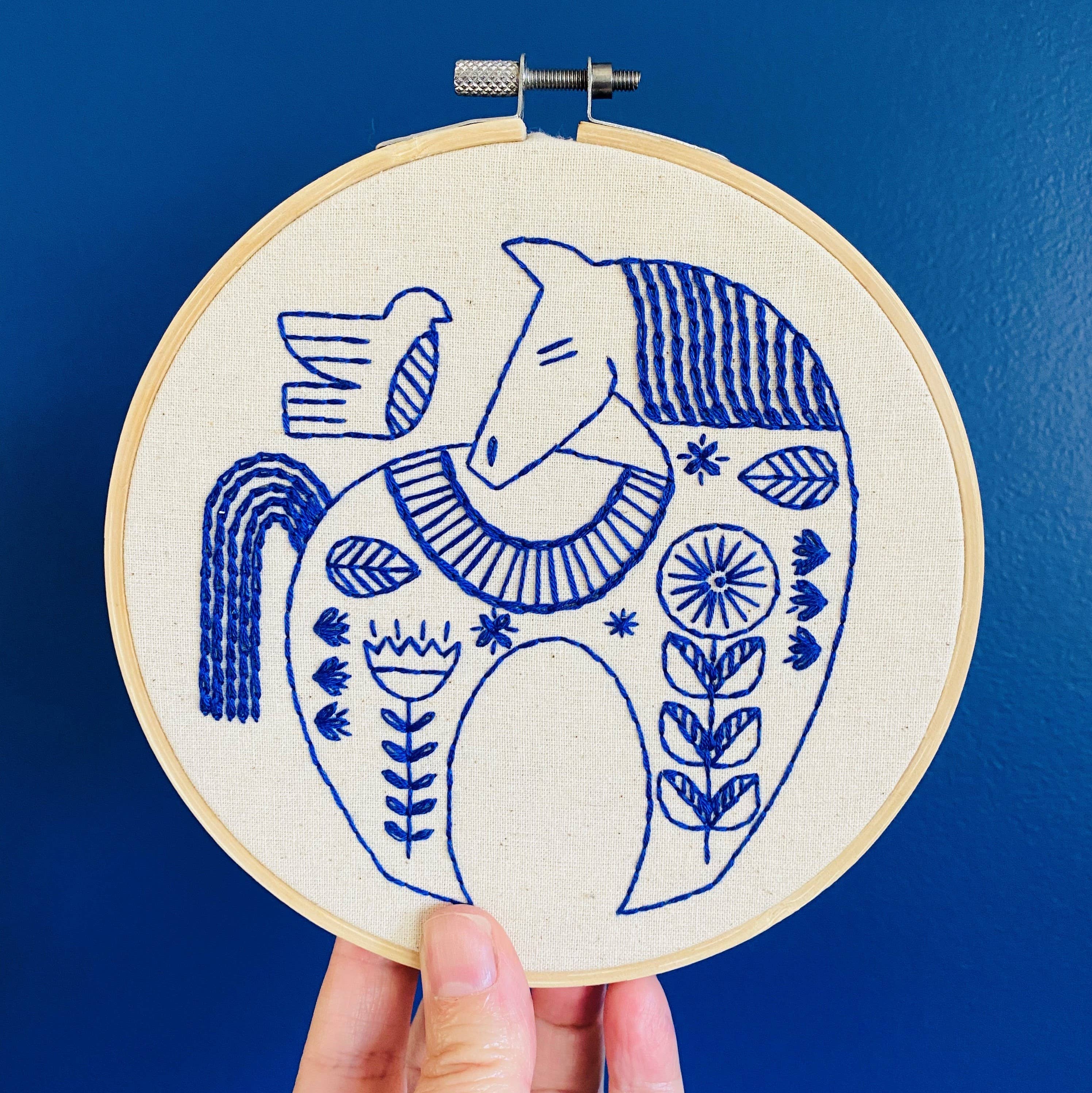 A hand holds up the Hygge Horse Complete Embroidery Kit by Hook, Line & Tinker, featuring a blue and white stylized horse, bird, and floral design stitched on cream fabric, set against a solid blue background.