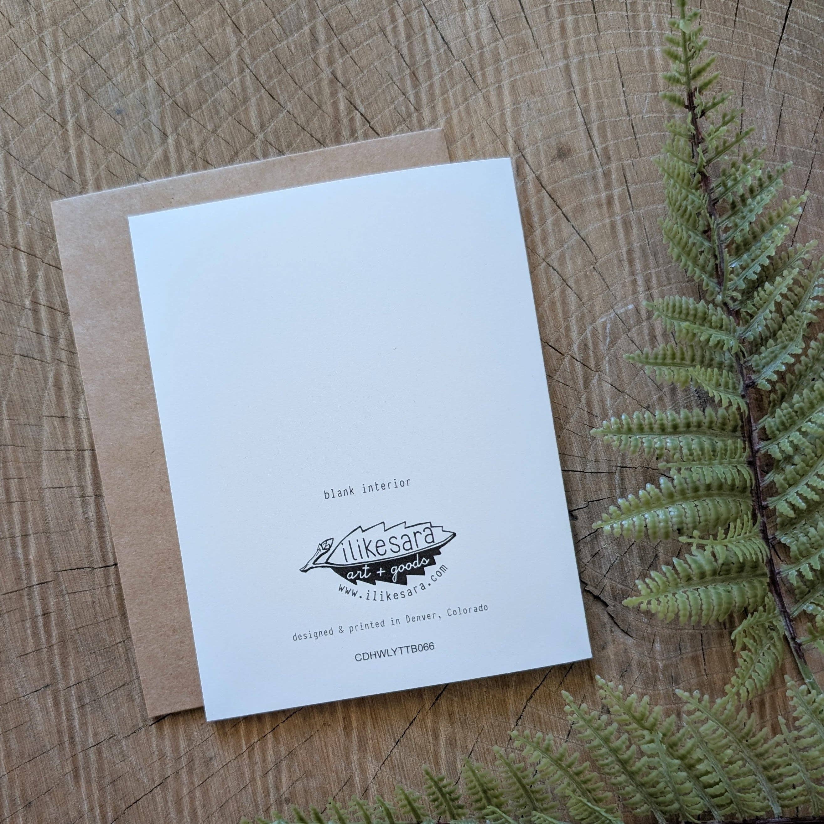 The "i love you to the bone" greeting card by ilikesara | art + goods rests on a wooden surface with a brown envelope and green fern. The blank white back shows the brand logo, website, and notes its blank interior.