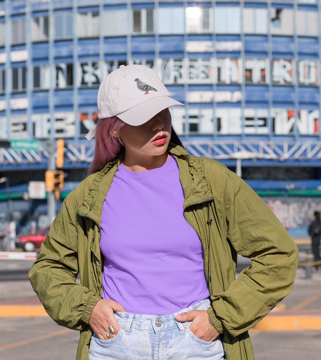 A person with pink hair wears a white cap decorated with the Rock Dove Pigeon, Birds, Iron on Patch by Wholesale Applique, paired with a green jacket and purple shirt, standing outdoors before a blue building with large windows and signage.