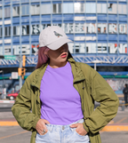 A person with pink hair wears a white cap decorated with the Rock Dove Pigeon, Birds, Iron on Patch by Wholesale Applique, paired with a green jacket and purple shirt, standing outdoors before a blue building with large windows and signage.