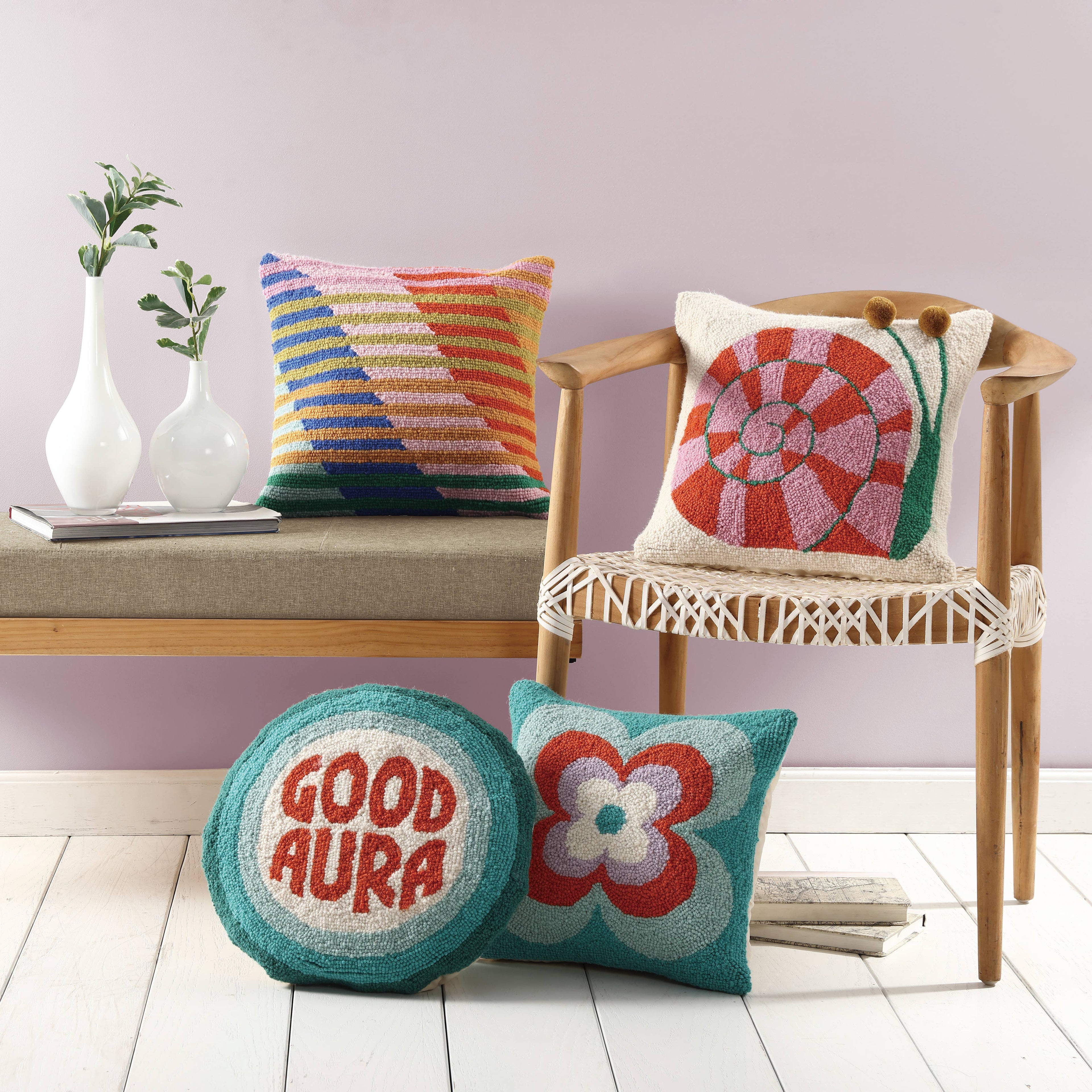 A modern room with a bench and chair features four colorful pillows—like the Snail's Pace w/Pom Pom Eyes Hook Pillow by Elizabeth Olwen for Peking Handicraft—plus white vases with green branches for a fresh, stylish look.