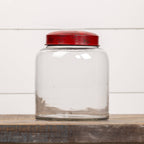 The Ragon House 8" CLEAR GLASS JAR W/ RED LID is shown empty on a wooden surface with a white paneled background.