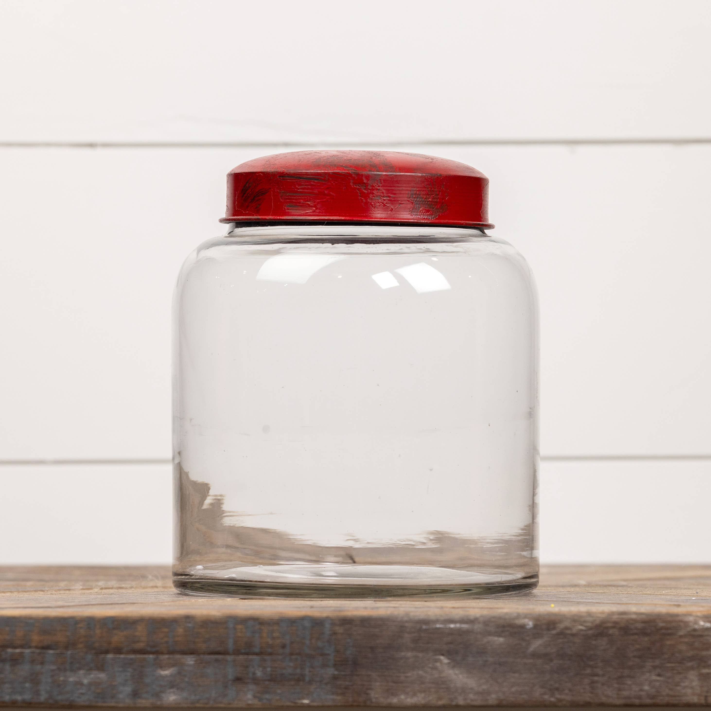 The Ragon House 8" CLEAR GLASS JAR W/ RED LID is shown empty on a wooden surface with a white paneled background.