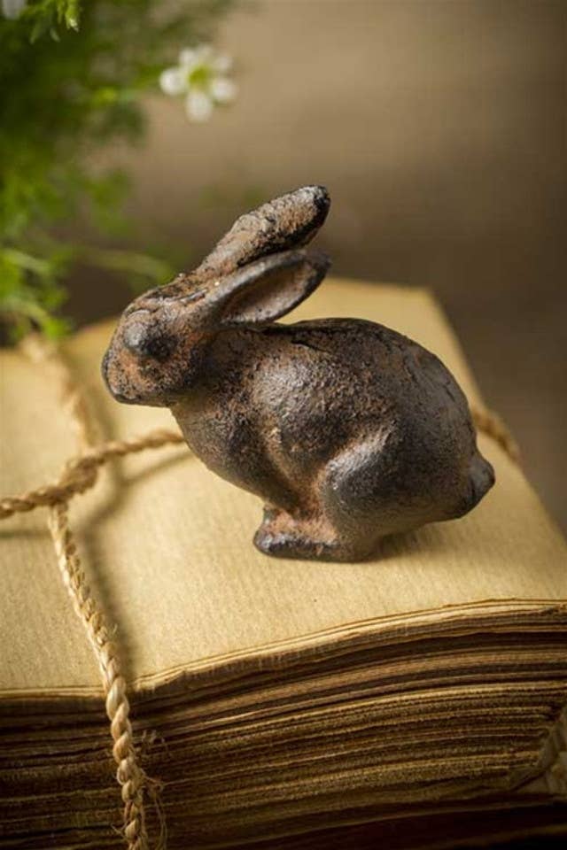 The Small Cast Iron Rabbit by Vagabond Vintage rests on aged papers tied with twine, with blurred greenery and white flowers in the background, adding timeless charm to this decorative accent.