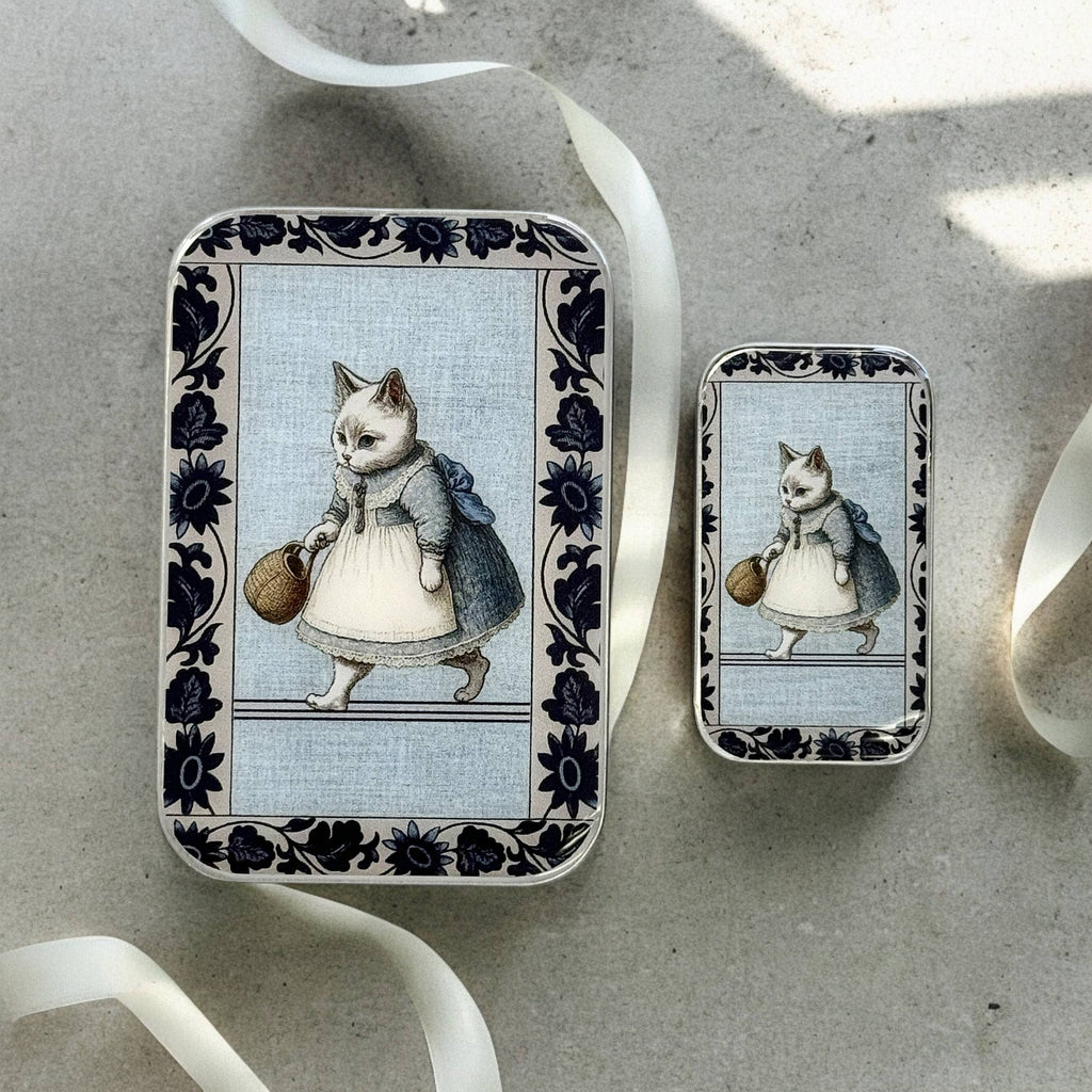 Two small Kitty notions tins by Firefly Notes feature vintage-style illustrations of a white "Business Cat" in a blue dress with basket, dark floral borders, and secure snap closure. Each rests on a light surface with white ribbon.