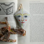 A Flying Kite ornament by Skippy Cotton, filled with cotton and lavender, lies on an open book. Shaped like a diamond kite, it features embroidery, red cheeks, a patterned tail—perfect as a scented gift topper.