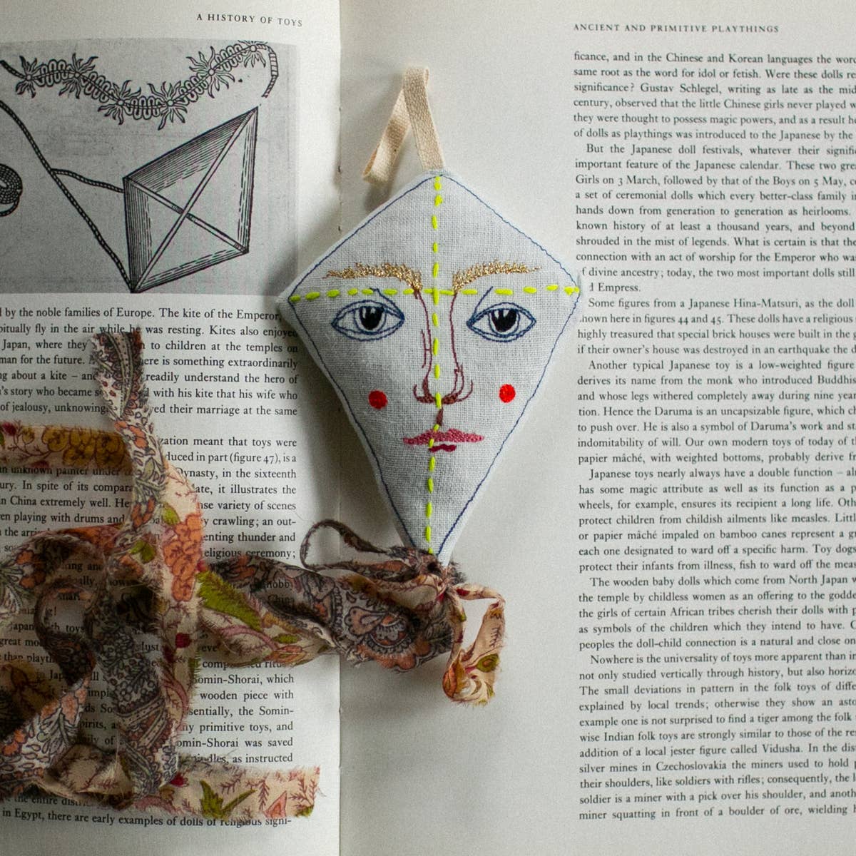 A Flying Kite ornament by Skippy Cotton, filled with cotton and lavender, lies on an open book. Shaped like a diamond kite, it features embroidery, red cheeks, a patterned tail—perfect as a scented gift topper.