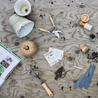 The Gardener's Planting Set by Bloomist, featuring gardening tools, floral scissors, seed packets, plant labels, string, and potted soil, rests on a wooden surface beside an open gardening book—perfect for planting prep.