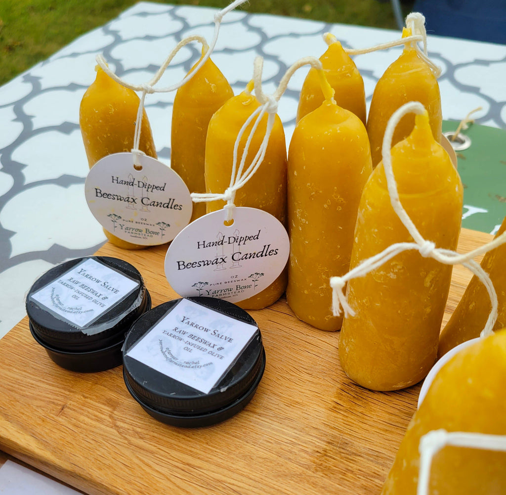 A display of Yarrow Bone Farmstead’s British Style Hand-dipped Beeswax Stubby Candles (pair), each with a tag, alongside tins of tallow salve arranged on a wooden board over a patterned tablecloth at an outdoor market.