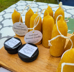 A display of Yarrow Bone Farmstead’s British Style Hand-dipped Beeswax Stubby Candles (pair), each with a tag, alongside tins of tallow salve arranged on a wooden board over a patterned tablecloth at an outdoor market.