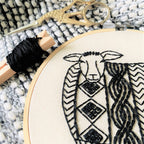 Close-up of black embroidery on beige fabric in a hoop, showing a stylized sheep with detailed wool. Ornate gold scissors and black thread rest on knit fabric—part of the Sweater Weather Sheep Embroidery Kit by Hook, Line & Tinker.