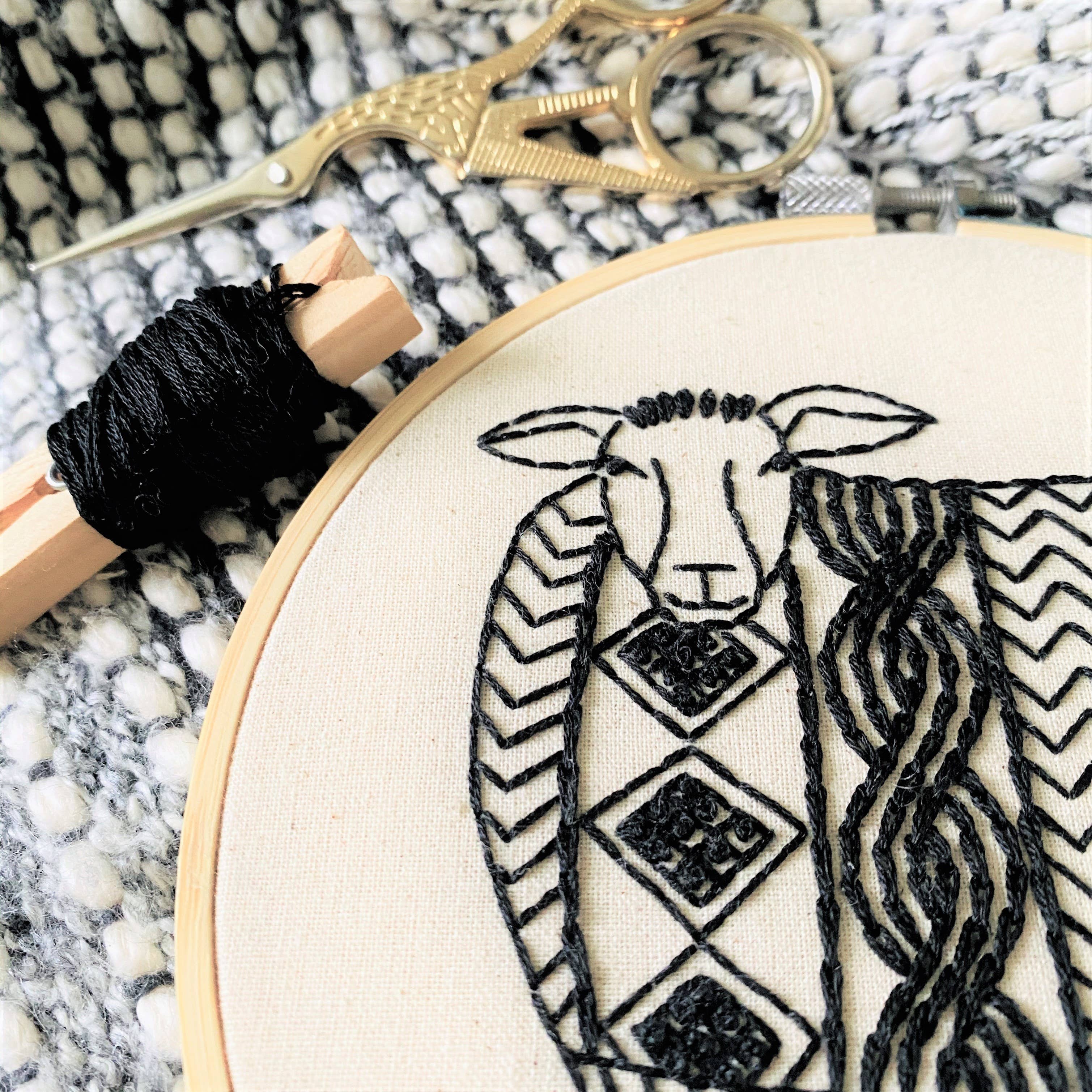 Close-up of black embroidery on beige fabric in a hoop, showing a stylized sheep with detailed wool. Ornate gold scissors and black thread rest on knit fabric—part of the Sweater Weather Sheep Embroidery Kit by Hook, Line & Tinker.