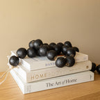The Foreside Home & Garden Black Terracotta Bead + Jute Garland rests atop three stacked books—Handmade, Homebody, and The Art of Home—on a wooden surface against a neutral wall.