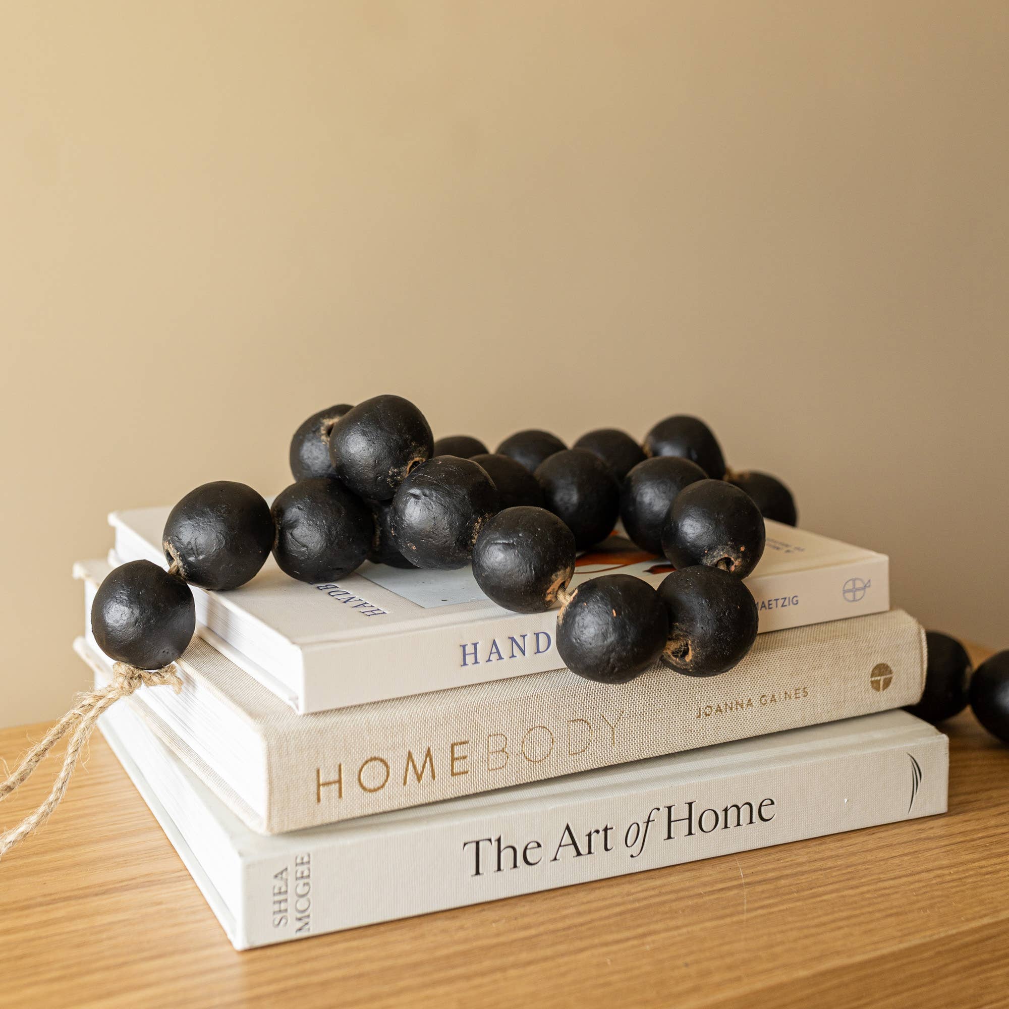 The Foreside Home & Garden Black Terracotta Bead + Jute Garland rests atop three stacked books—Handmade, Homebody, and The Art of Home—on a wooden surface against a neutral wall.