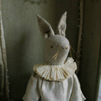 The Skippy Cotton Ruffle Bun Ragdoll is a cotton-filled art doll in a charming cotton dress, with long ears, embroidered face, ruffled collar, and delicate hand-stitched details, set against a rustic background.