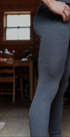 Someone wearing Choice Alpaca Products' Alpaca Leggings, featuring a gray textured, moisture-wicking design, stands indoors by a wooden table and chair. Natural light streams through a window, highlighting only the lower torso and legs.