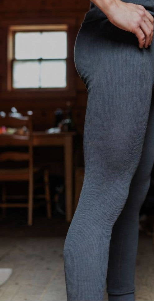 Someone wearing Choice Alpaca Products' Alpaca Leggings, featuring a gray textured, moisture-wicking design, stands indoors by a wooden table and chair. Natural light streams through a window, highlighting only the lower torso and legs.