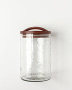 The Handblown Hammered Glass Canisters: Smoke / Small by Creative Women features a textured surface, airtight seal, and hand-carved wooden lid, displayed against a plain white background.