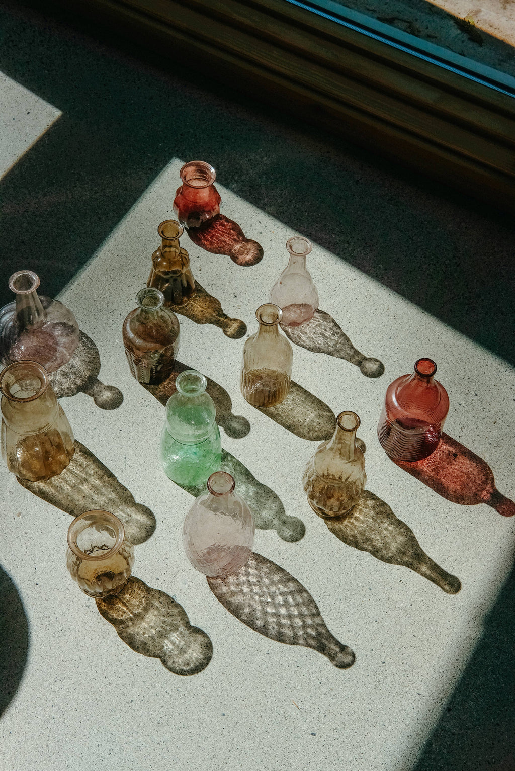 The Indaba Trading Recycled Glass Vase in Sangria sits on a sunlit surface, casting vibrant pink and brown shadows as light streams through a nearby window.