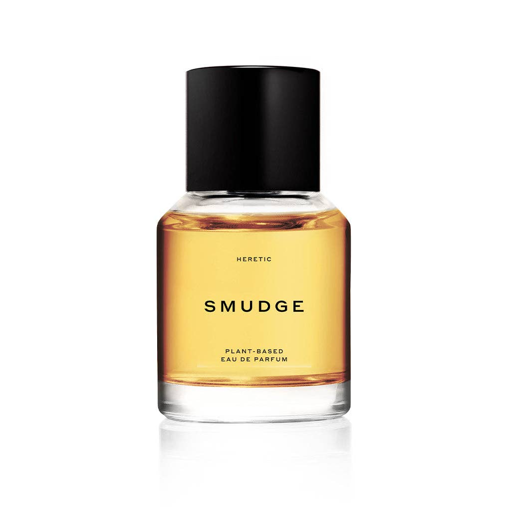 A clear glass bottle of Heretic Parfum’s SMUDGE: 15ML, filled with amber liquid and topped with a black cylindrical cap, displays "PLANT-BASED EAU DE PARFUM" on its label and evokes frankincense notes against a crisp white background.