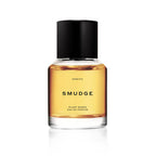 A clear glass bottle of Heretic Parfum’s SMUDGE: 15ML, filled with amber liquid and topped with a black cylindrical cap, displays "PLANT-BASED EAU DE PARFUM" on its label and evokes frankincense notes against a crisp white background.