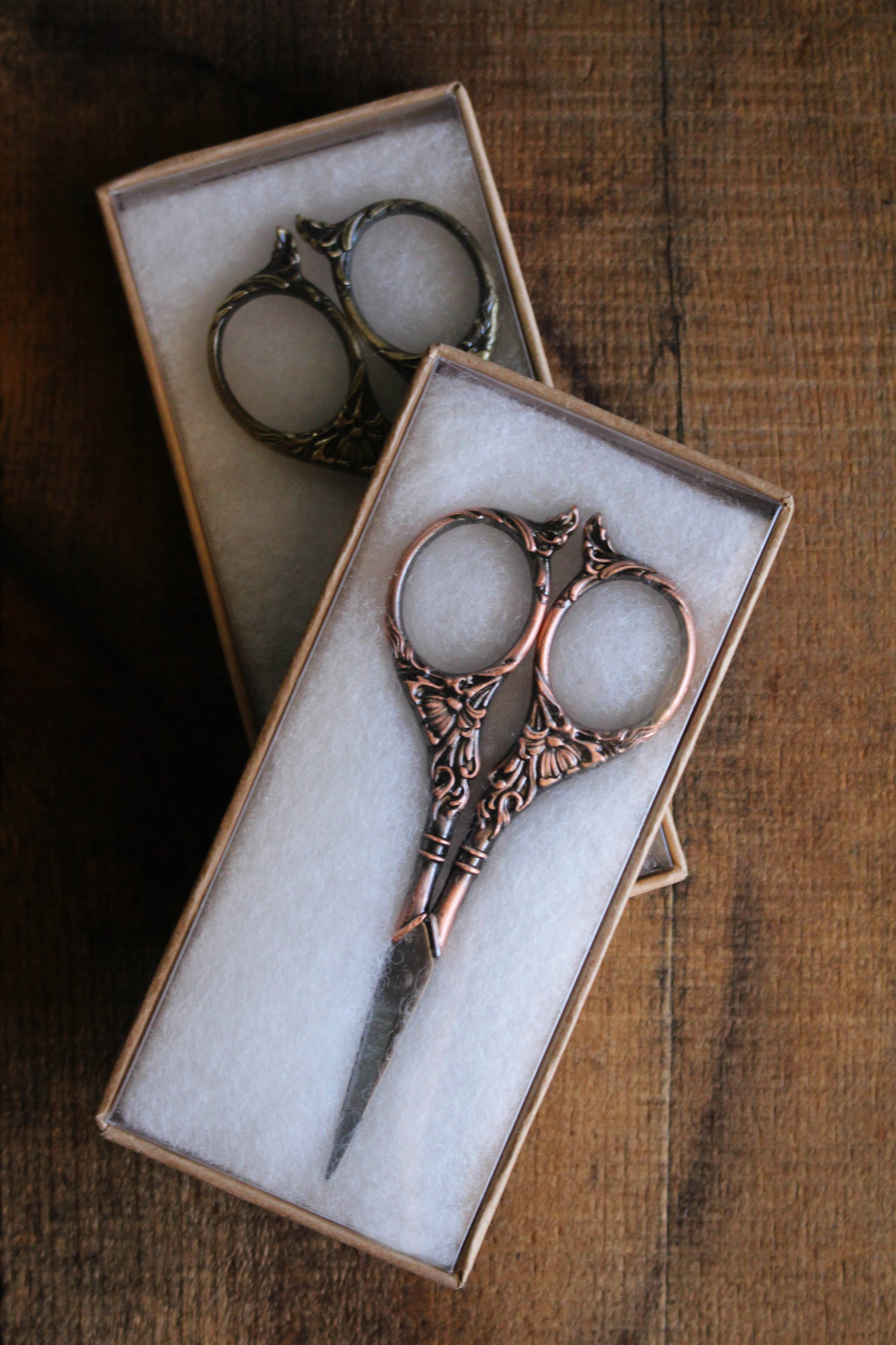 The NNK Press Botanical Garden Scissors in Antique Gold features ornate, botanical-inspired handles and comes in a cream-lined box displayed on a wooden surface.