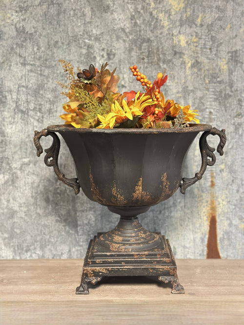 The Vagabond Vintage Distressed Black Iron Urn with lion head handles holds yellow, orange, and brown artificial flowers, displayed on wood with a textured gray backdrop.