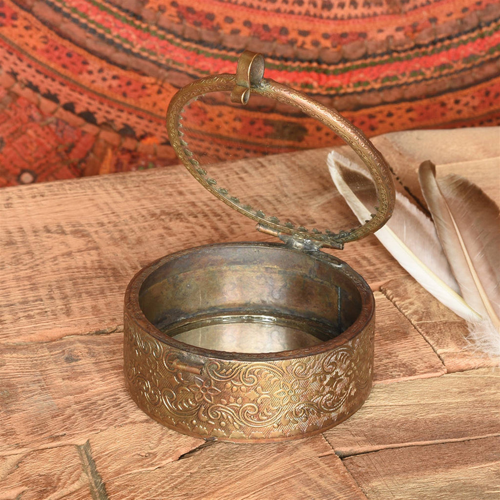 The HomArt Josephine Mirrored Box, Brass - Sm Round, featuring intricate floral engravings and a glass lid, rests open on wood near two feathers with a patterned reddish textile in the background.