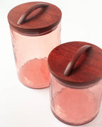 Two Handblown Hammered Glass Canisters: Blush / Small by Creative Women, featuring textured pink glass and hand-carved wooden lids with curved handles, are displayed on a white background.
