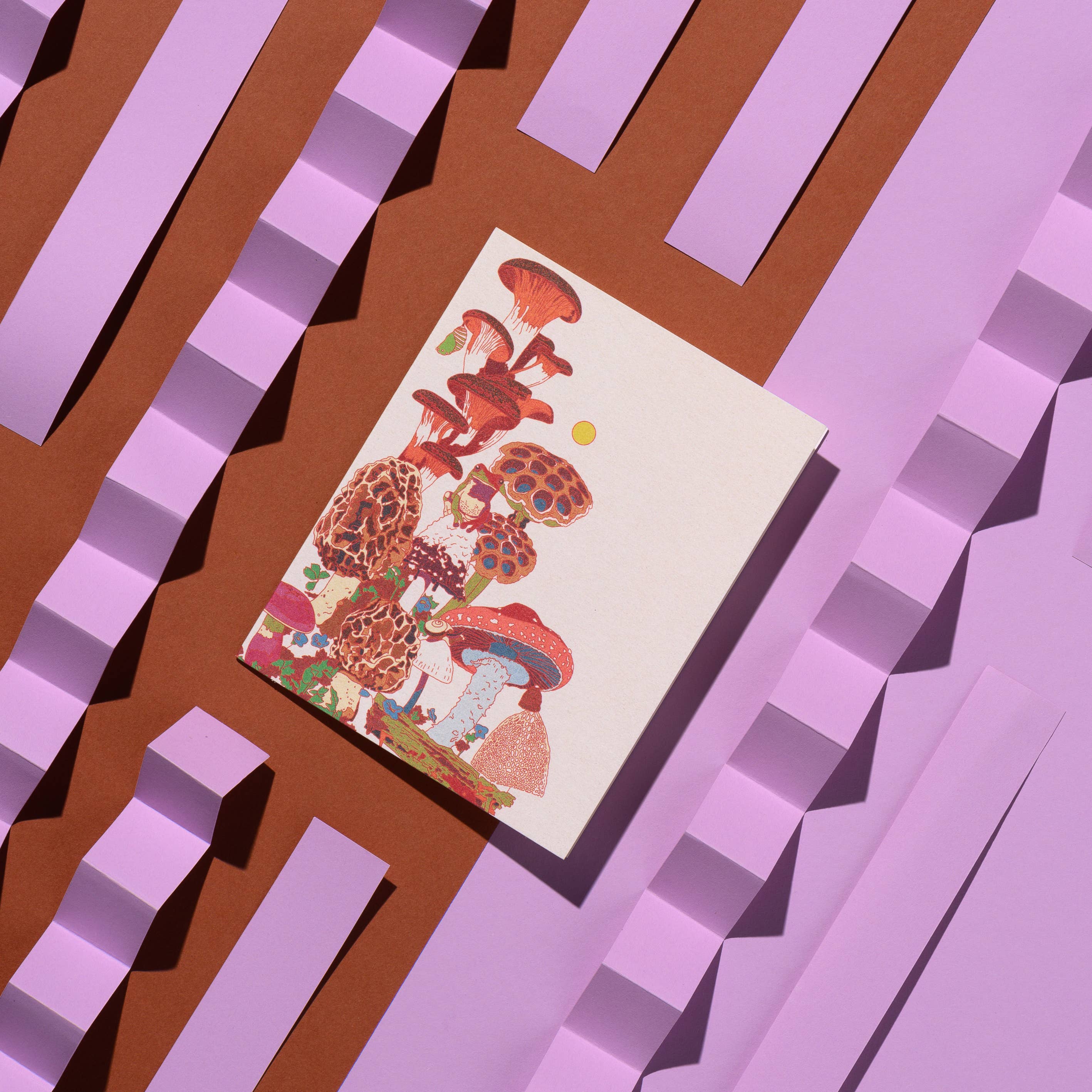 The Frog and Fungi greeting card: Single by Red Cap Cards features colorful illustrated mushrooms and is displayed on a brown surface with layered, folded pastel purple paper strips in a geometric design.