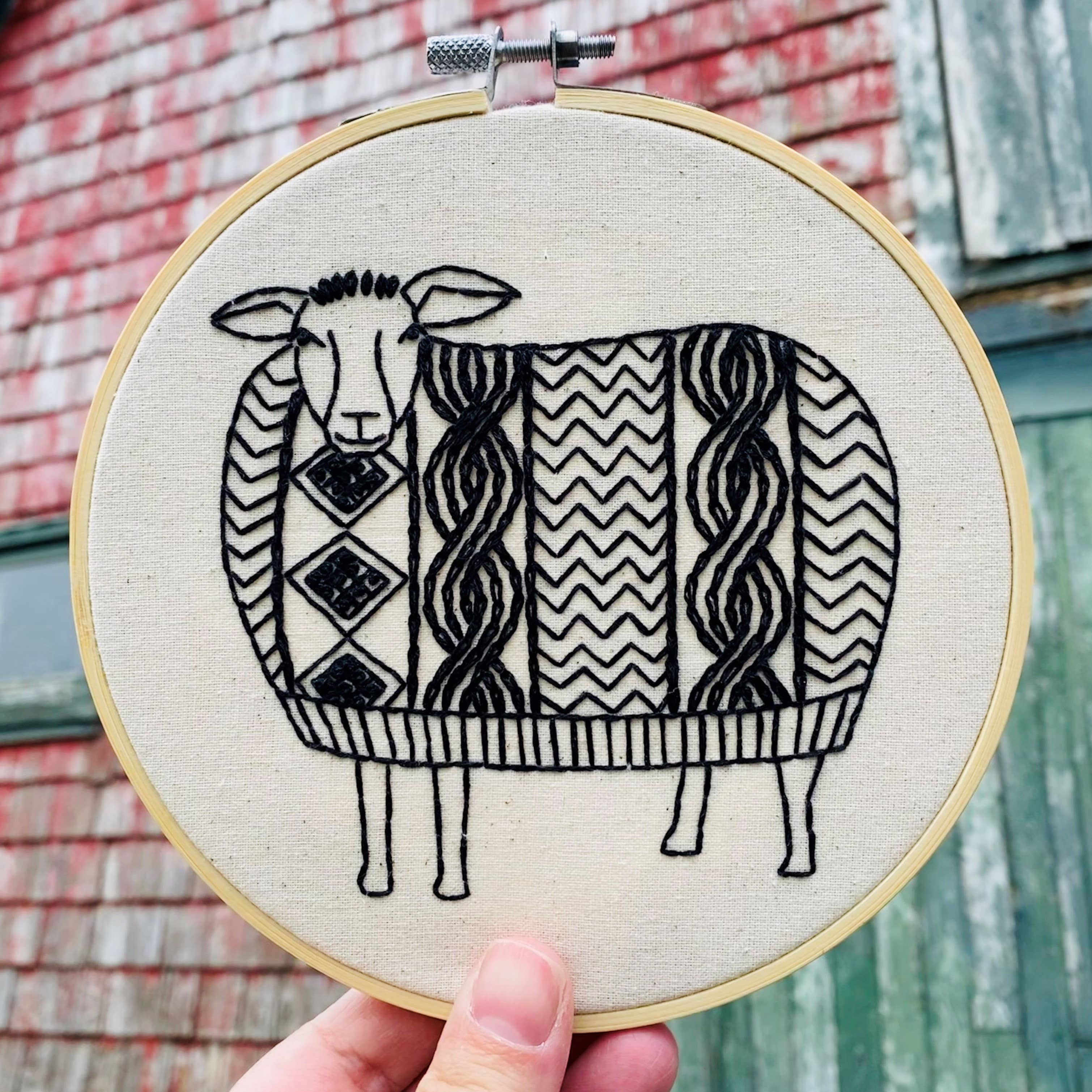 A hand displays the Sweater Weather Sheep Complete Embroidery Kit by Hook, Line & Tinker Embroidery Kits, featuring a black embroidered sheep with a patterned wool coat against a backdrop of rustic, weathered buildings.