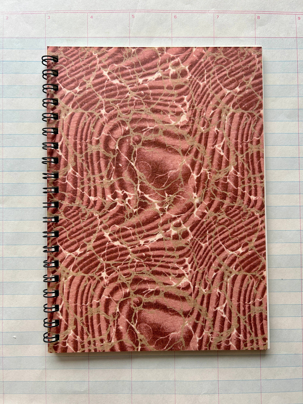 The Parcel Antique Endpaper Notebook: Small - Composition / Unlined, featuring a textured marbled brown and pink cover, sits atop a 5x7 lined paper with blue and pink grid lines.