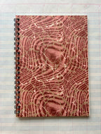 The Parcel Antique Endpaper Notebook: Small - Composition / Unlined, featuring a textured marbled brown and pink cover, sits atop a 5x7 lined paper with blue and pink grid lines.