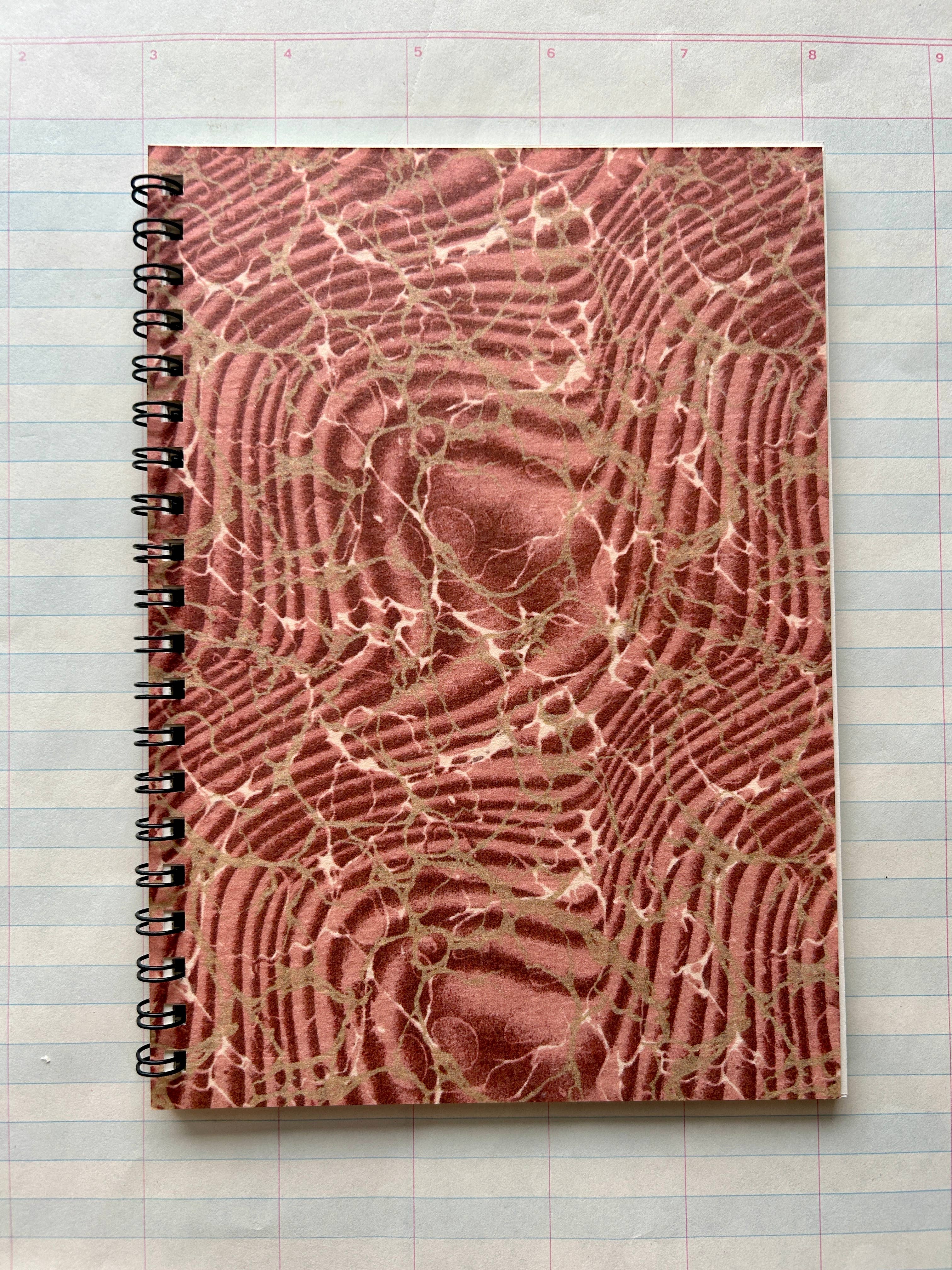 The Parcel Antique Endpaper Notebook: Small - Composition / Unlined, featuring a textured marbled brown and pink cover, sits atop a 5x7 lined paper with blue and pink grid lines.