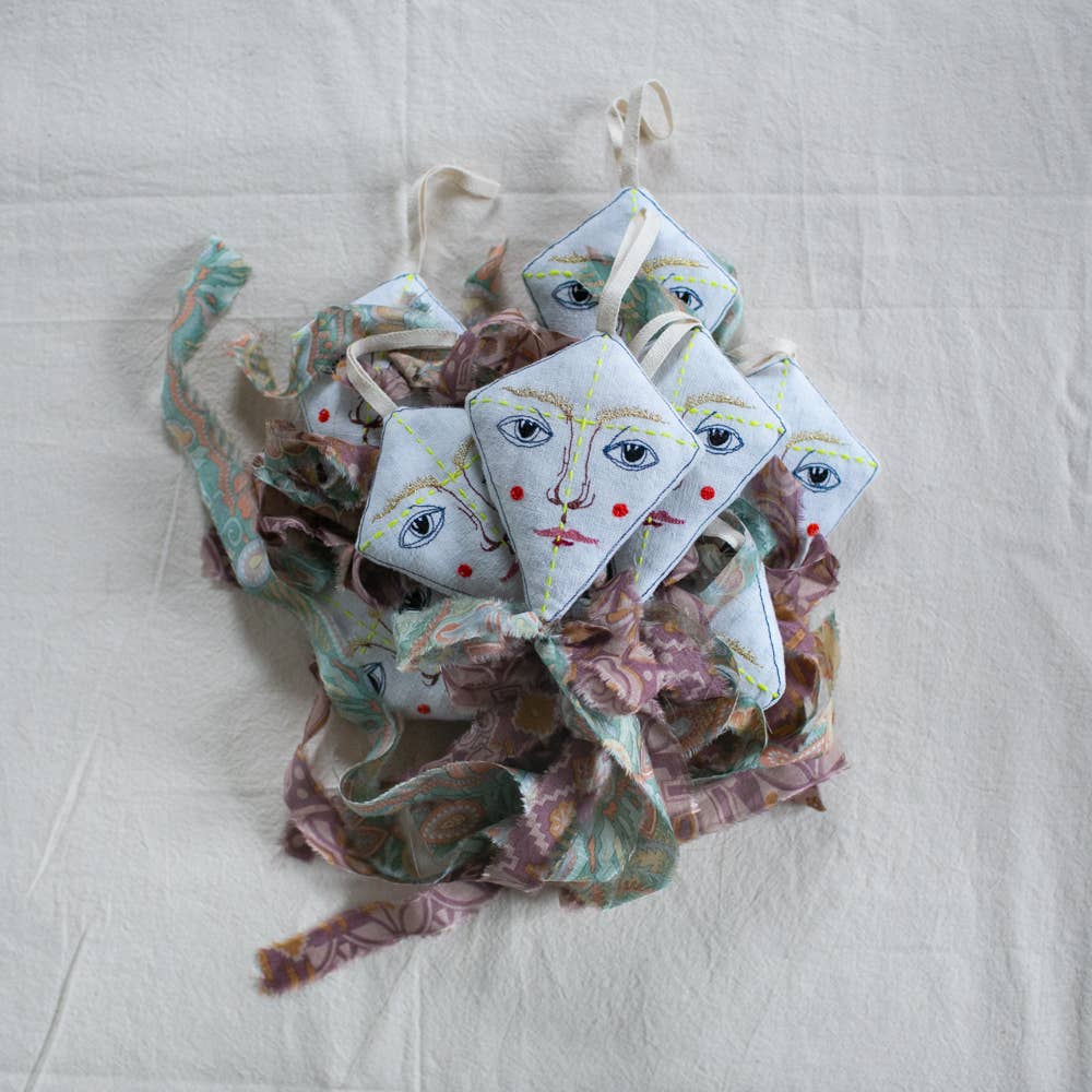 The Flying Kite Cotton & Lavender filled Ornament by Skippy Cotton features diamond-shaped, handmade organic cotton designs with embroidered faces, yellow lines, and colorful ribbon details, beautifully displayed on a white cloth. Scented for added charm.