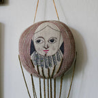The Skippy Cotton Wonderful Wizard / Balloon Oz wallhanging ornament features an embroidered face with long hair, red lips, a detailed collar, and hangs from gold beads against a white wall.