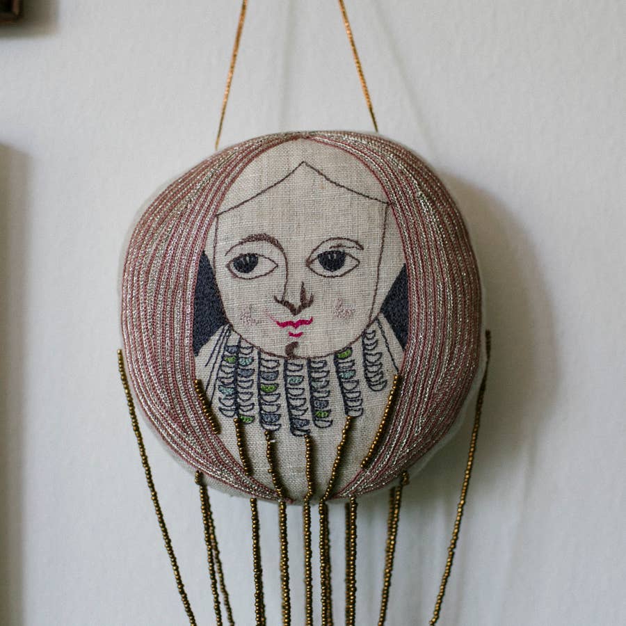 The Skippy Cotton Wonderful Wizard / Balloon Oz wallhanging ornament features an embroidered face with long hair, red lips, a detailed collar, and hangs from gold beads against a white wall.
