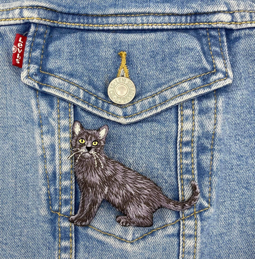 The Wholesale Applique Black Cat Realistic Full Body Embroidered Iron on Patch features yellow eyes and detailed stitching, perfect for customizing blue denim jackets or other outerwear with a unique pet-themed accent.