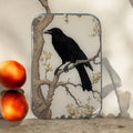 A framed illustration of a black raven on a branch leans against a wall. Next to it, two peaches and the Firefly Notes Black Crow Notions Tin – Handcrafted form an artful display on the light surface.