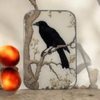 A framed illustration of a black raven on a branch leans against a wall. Next to it, two peaches and the Firefly Notes Black Crow Notions Tin – Handcrafted form an artful display on the light surface.