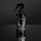 A black 200ml O TANNENBAUM ROOM SPRAY by Heretic Parfum, featuring Edward Gorey-style white art of a child, trees, and a dog on the label, sits on a reflective black surface—hinting at its unique fir needle fragrance.