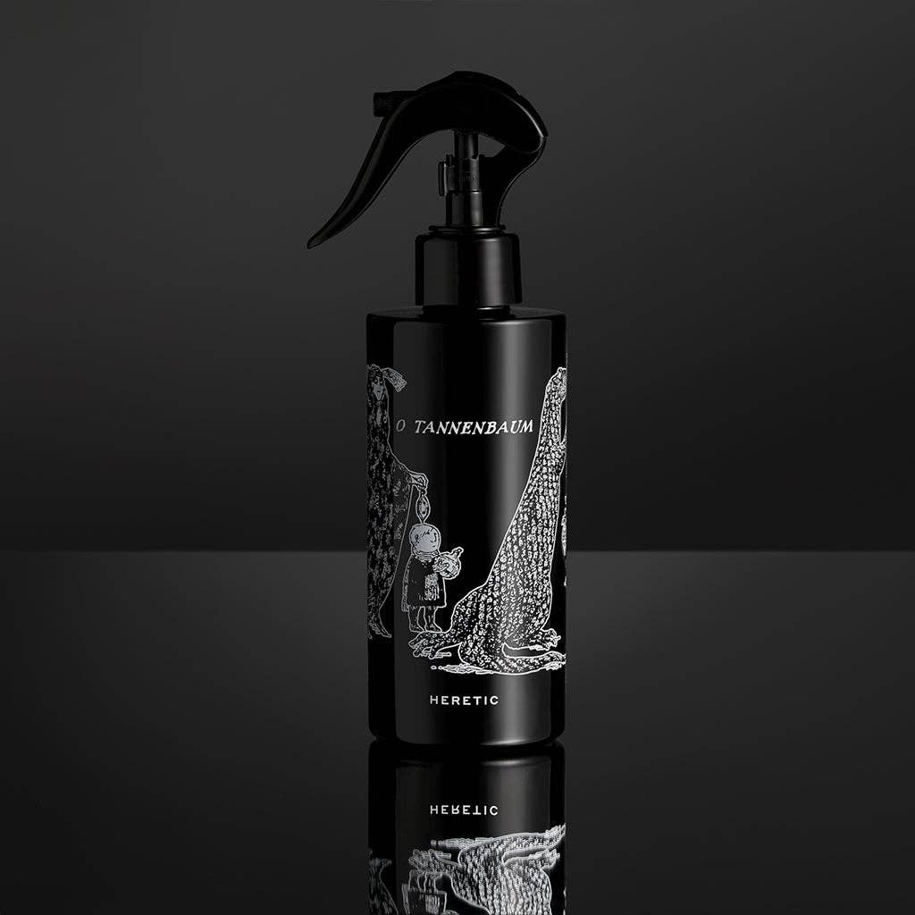 A black 200ml O TANNENBAUM ROOM SPRAY by Heretic Parfum, featuring Edward Gorey-style white art of a child, trees, and a dog on the label, sits on a reflective black surface—hinting at its unique fir needle fragrance.
