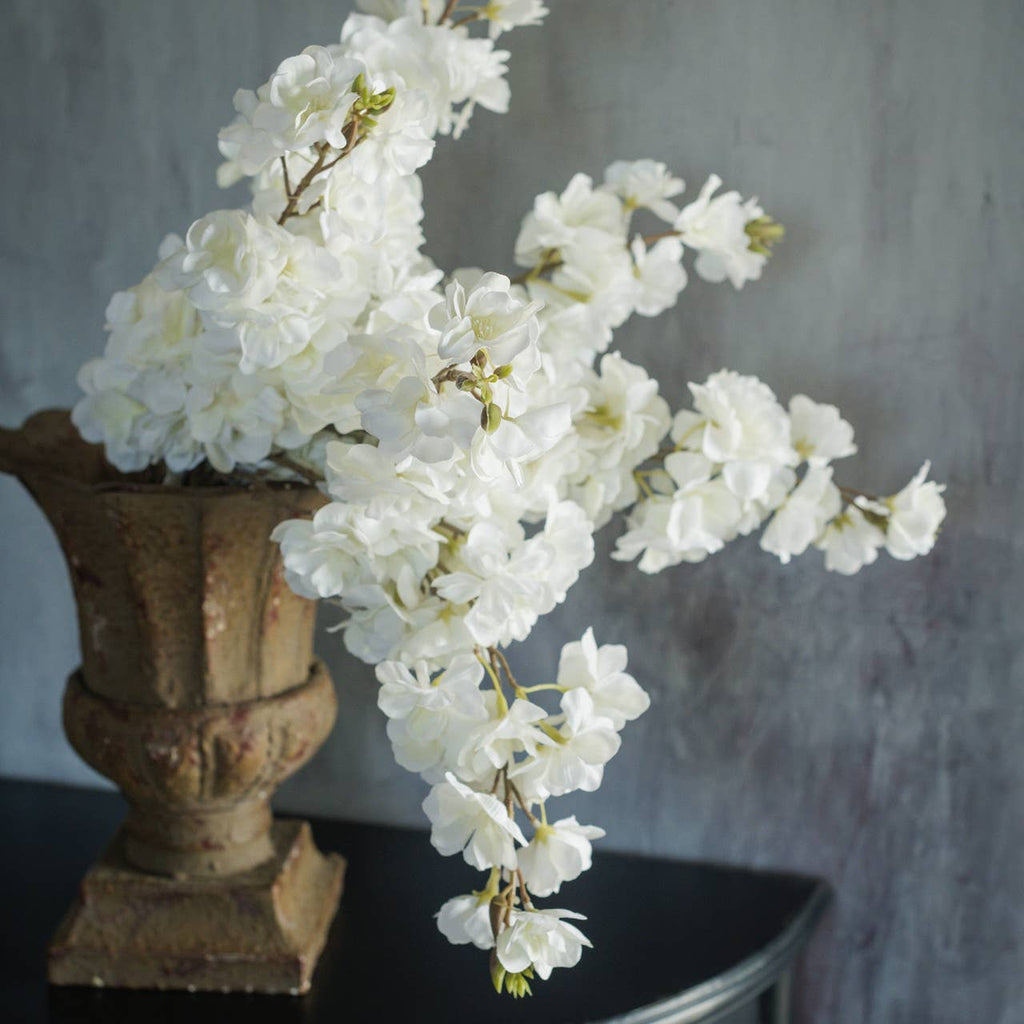 A Ragon House FAUX WHITE CHERRY BLOSSOM SPRAY elegantly cascades from a rustic brown vase on a dark table, set against a textured gray backdrop for a refined faux floral arrangement.