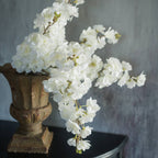 A Ragon House FAUX WHITE CHERRY BLOSSOM SPRAY elegantly cascades from a rustic brown vase on a dark table, set against a textured gray backdrop for a refined faux floral arrangement.